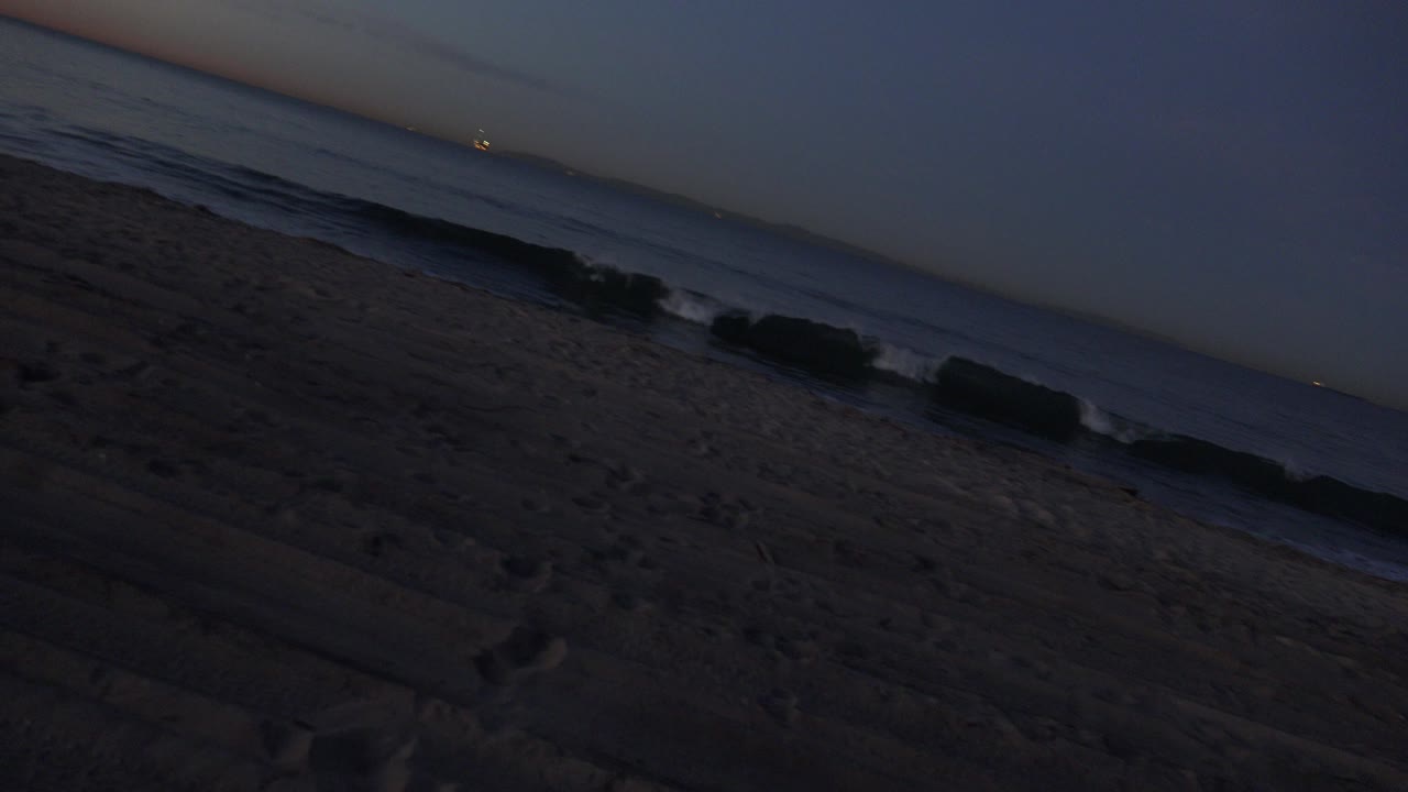Huntington Beach shoreline at dusk with dark sand in the foreground, small waves rolling in, a thin warm horizon glow, and no visible people.