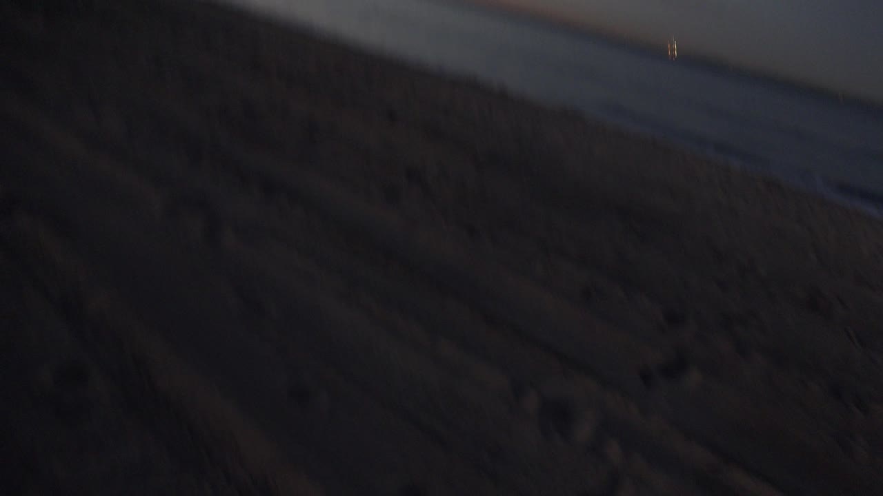 Tilted opening view of Huntington Beach at dusk, with dark footprint-marked sand in front, the ocean to the right, an orange sunset band on the horizon, and no visible people.