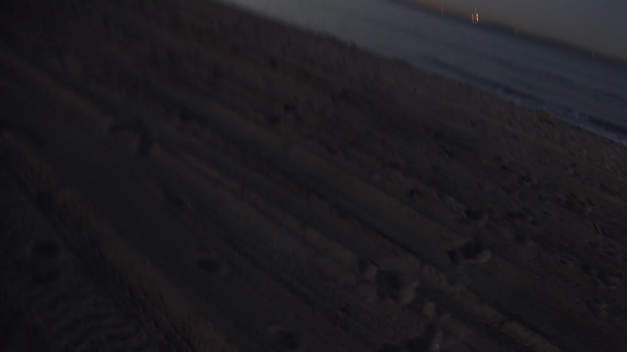 Huntington Beach shoreline at dusk, with the sandy foreground mostly in shadow, low surf along the right edge, blue sky above an orange horizon, and no visible people.