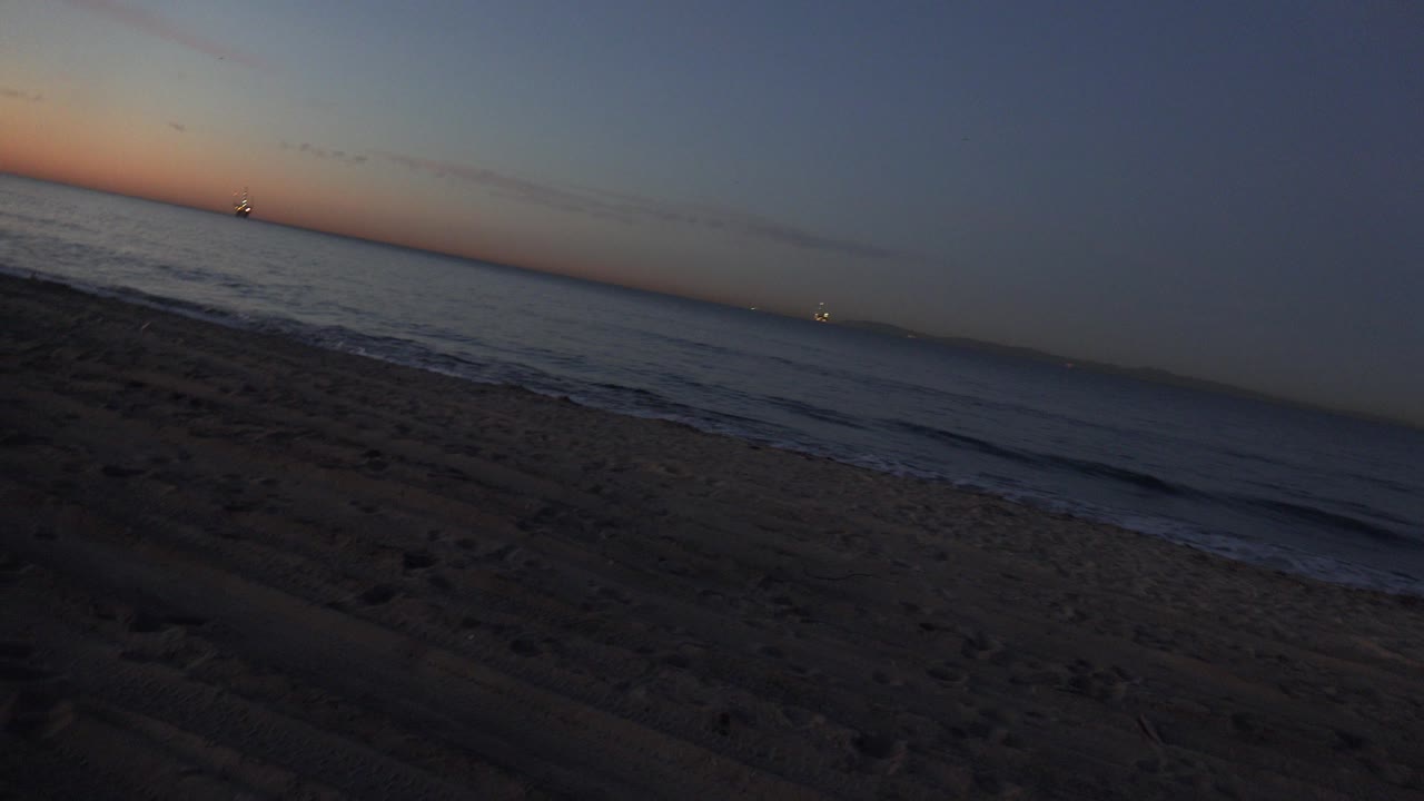 Opening frame of Huntington Beach at evening, with tan sand in the foreground, small blue-gray waves near shore, a tilted horizon, distant lights, and no visible people.