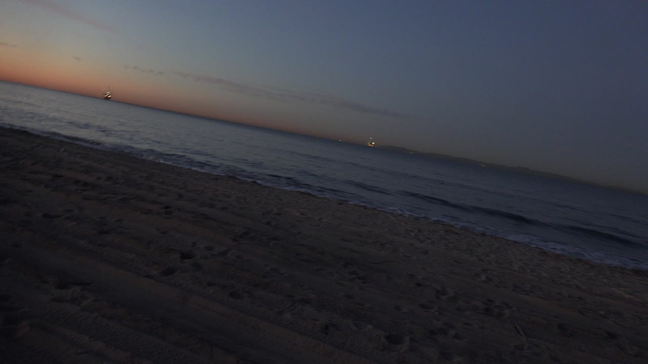 Huntington Beach shoreline one second in, showing darkened sand, low surf, a warm strip of sunset color near the horizon, distant lights, and no visible people.