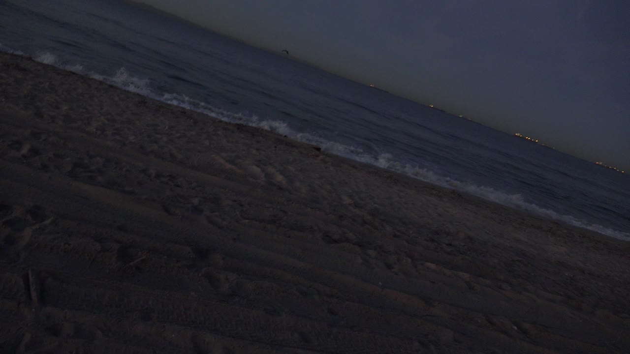 At 0:00, a tilted Huntington Beach dusk view shows dark textured sand below a thin breaking wave line and open ocean, with no visible people.