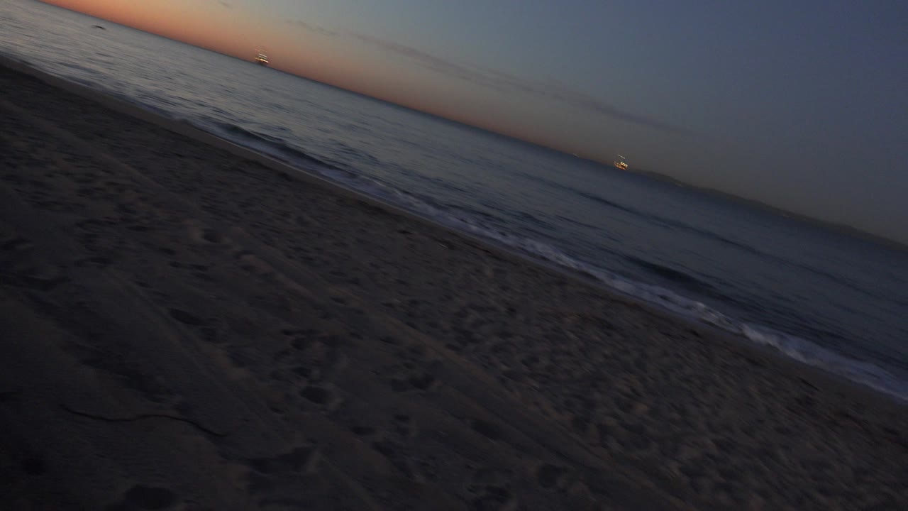 Tilted Huntington Beach view with dark sand in the foreground, surf along the shoreline, orange sunset light near the horizon, and no visible people.