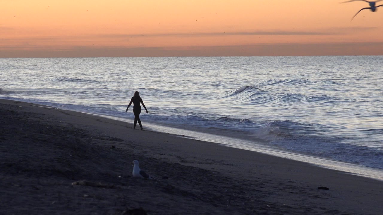 At Huntington Beach, California, a distant silhouetted person walks near the waterline as a small wave breaks under an orange sunset sky.