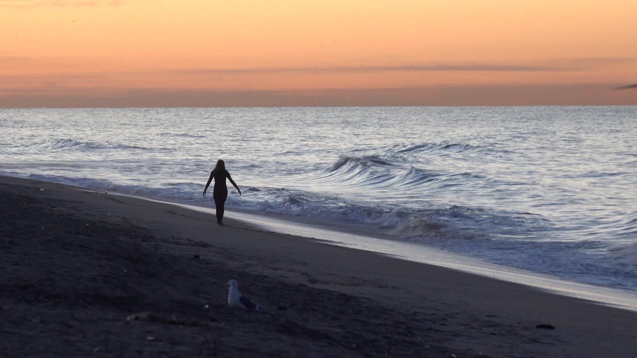 Lone beach walker approaching the waterline as waves roll in under sunset light.