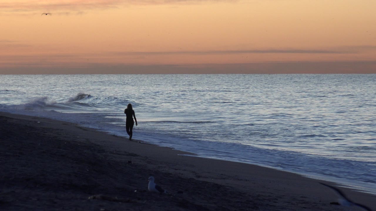The Huntington Beach shoreline remains wide and quiet as the silhouetted person stands farther along the wet sand while small waves roll in under the orange-blue sky.