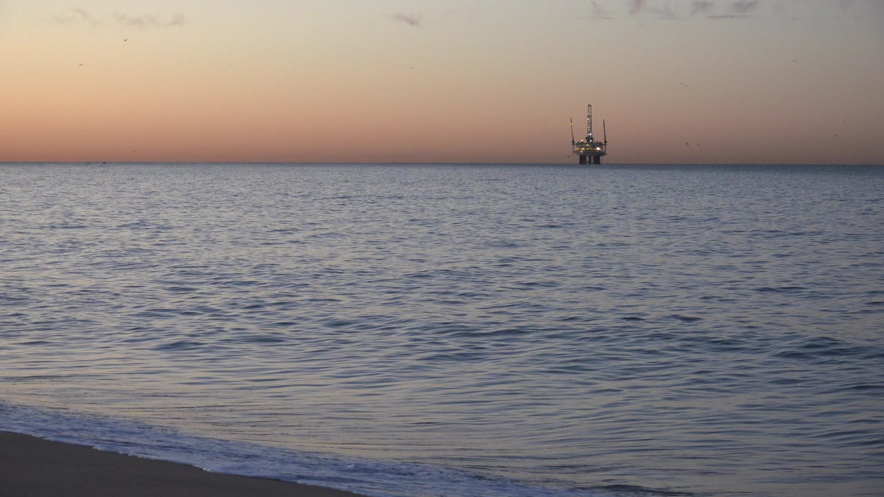 Huntington Beach ocean at pale pink light, with small waves reaching the dark sand and an offshore platform near the horizon, no visible people.
