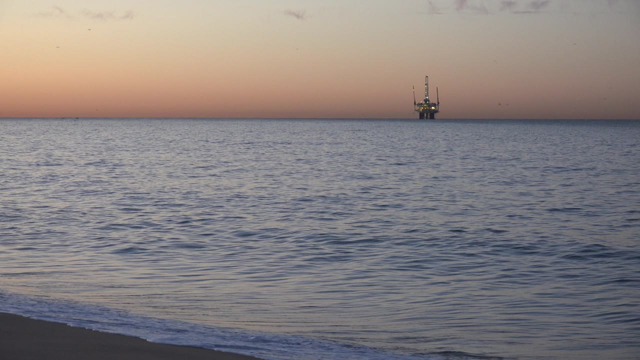 Huntington Beach shoreline with rippled water in the foreground, a peach-pink band above the horizon, and the offshore platform held small in the distance, no visible people.