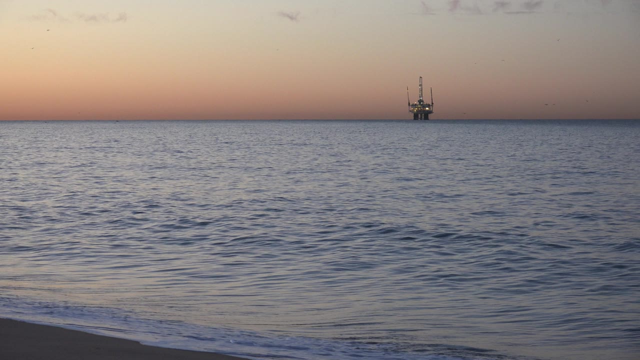 Gentle shore break in front of open ocean and a small offshore platform.