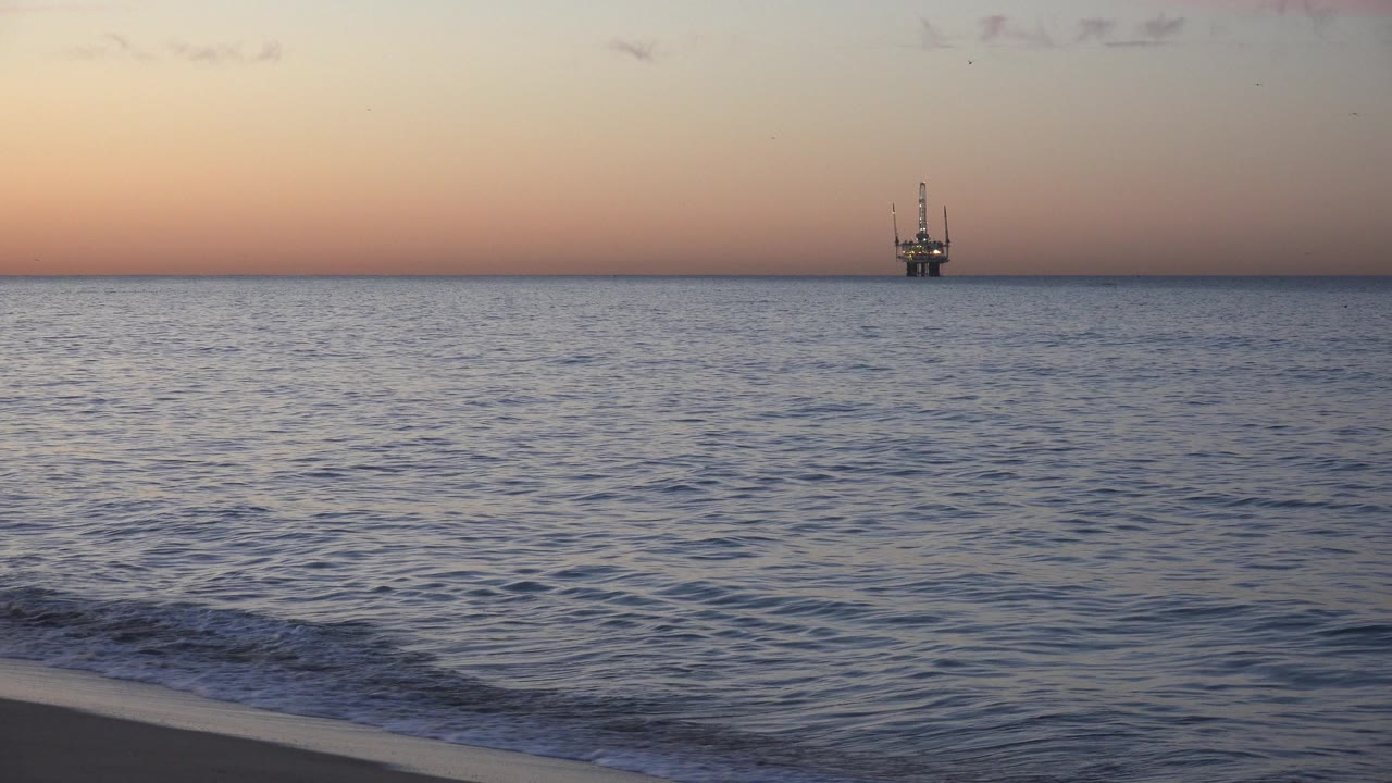 Foam spreads along the lower shoreline as the Huntington Beach ocean holds a distant vessel against a peach and blue dusk sky, with no visible people.