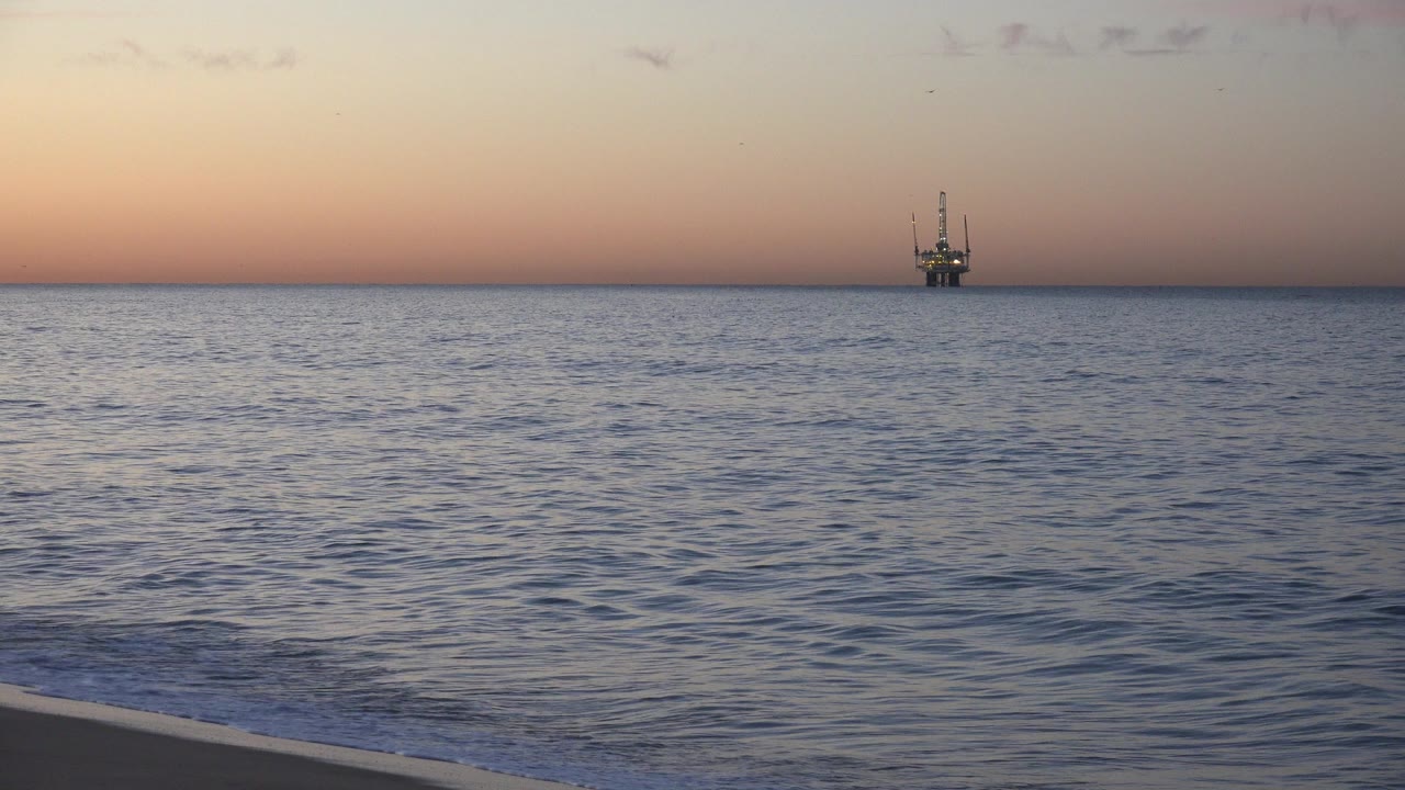 The Huntington Beach shoreline sits low in frame, calm rippled water fills the midground, and the offshore vessel appears near the horizon with no visible people.