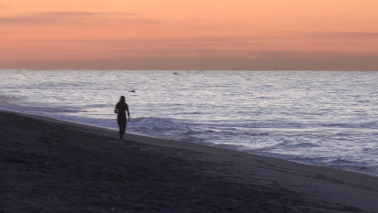 A silhouetted woman walks along the sloped sand at Huntington Beach, with small surf rolling in and a peach sunset sky above the horizon.