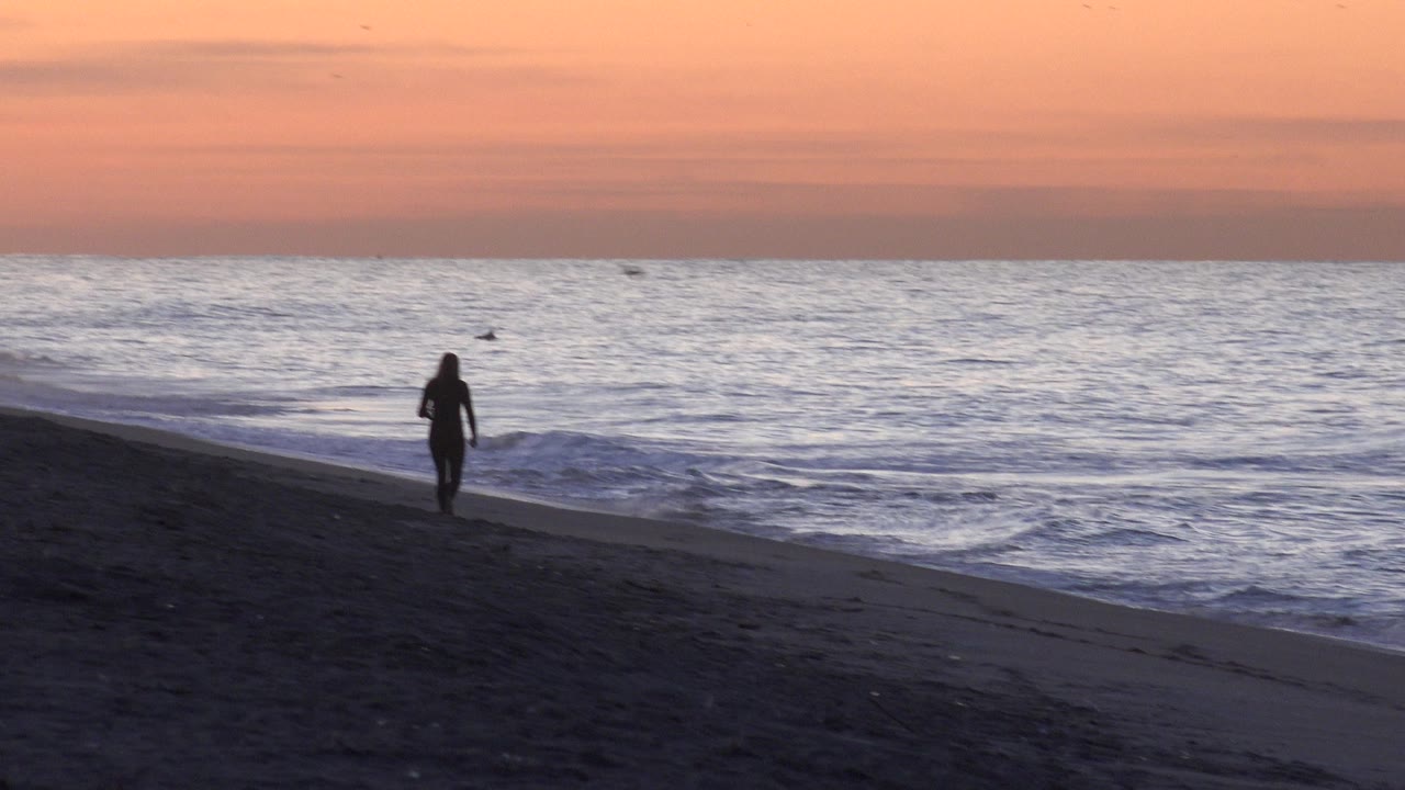 The woman remains in silhouette near the waterline at Huntington Beach as the gray blue ocean fills the background under warm dusk color.