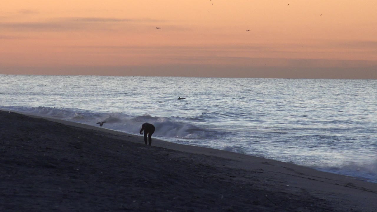 A silhouetted person bends or leans near the Huntington Beach shoreline as small waves break under an orange evening sky.
