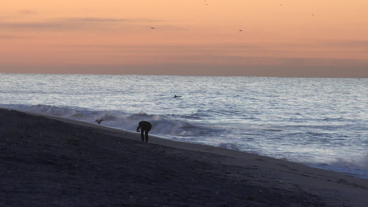 A dark figure stands or bends near the Huntington Beach surf line while a small wave curls in from the right beneath a peach-colored sky.