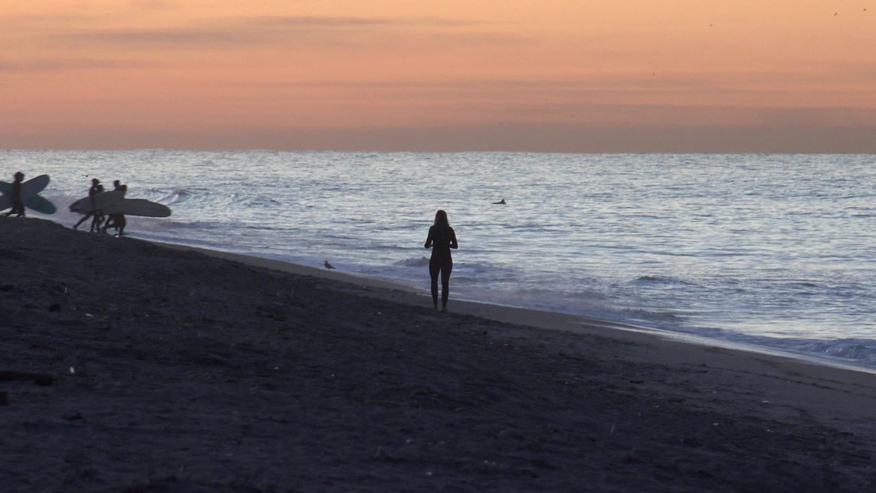 At Huntington Beach, California, a single silhouetted figure stands near the waterline while a small group with surfboards gathers farther left.