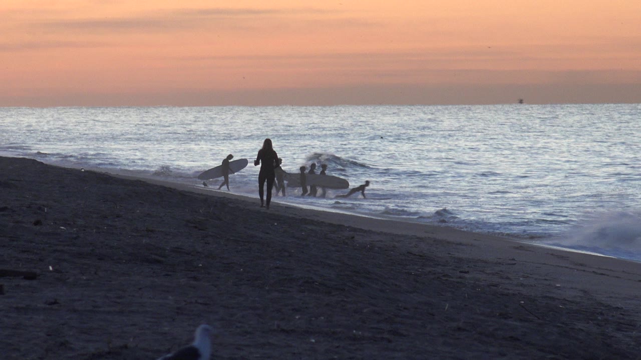 At Huntington Beach, California, one surfer stands near center while others hold surfboards at the shore and a low wave rolls toward the dark sand.