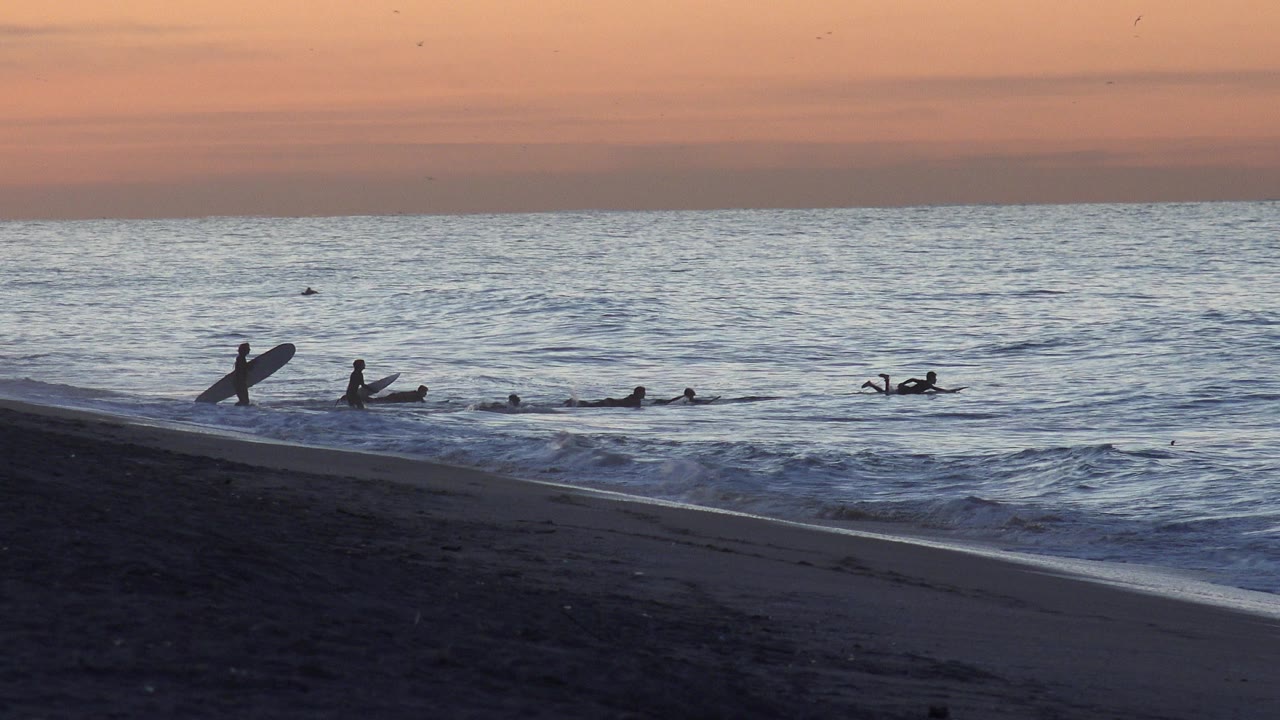A silhouetted surfer with a board stands near the left shoreline at Huntington Beach while several others sit or paddle beyond the small breaking waves.