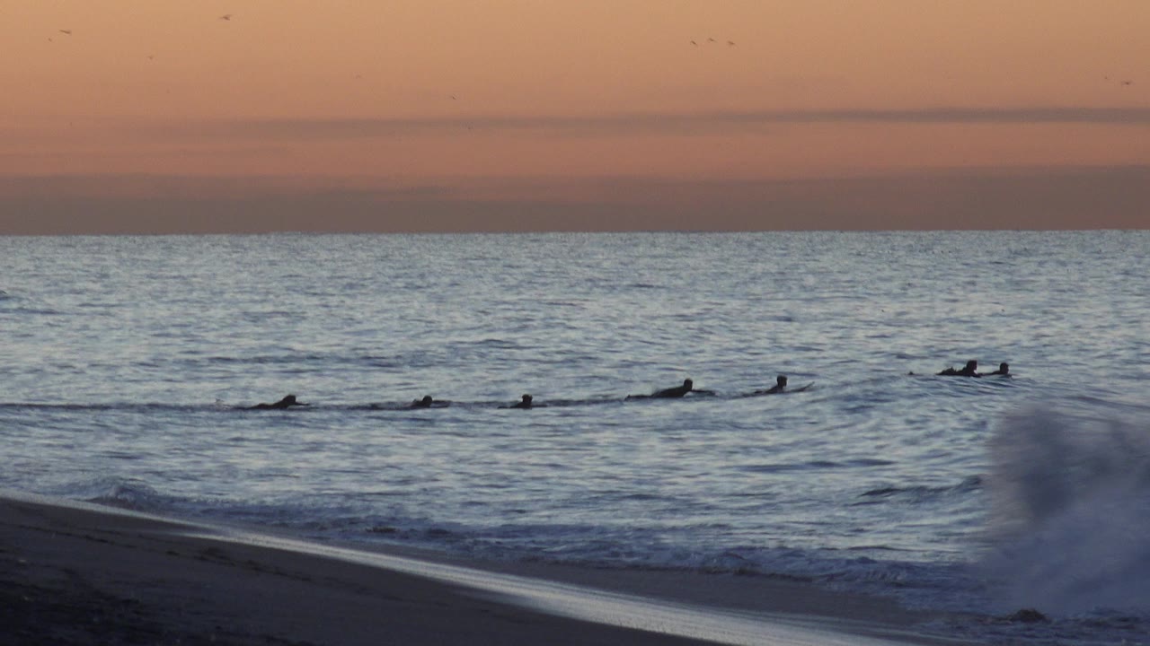 Distant surfers paddle offshore as a wave breaks near the beach at sunset.