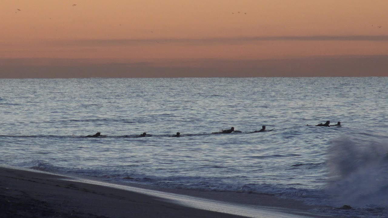Surfers remain spread across the midground at Huntington Beach, California, while a foamy wave rises in the foreground under a muted orange sky.
