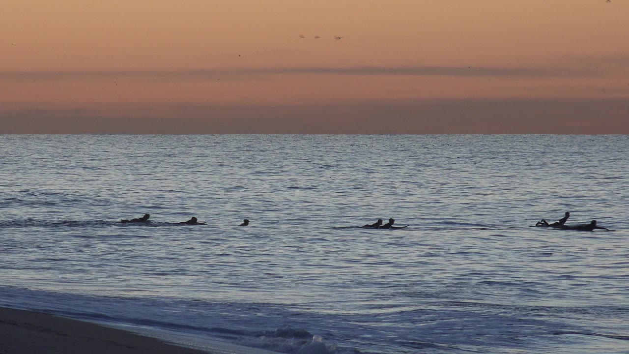 Seven distant surfers float and paddle in the ocean off Huntington Beach, California, with a low orange sky and small waves near shore.