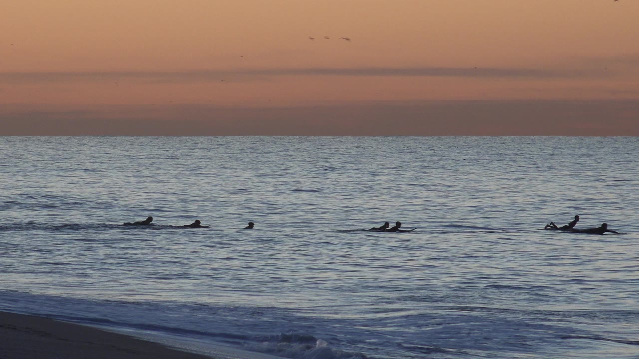 Distant surfers remain spread across the Huntington Beach water as a small foreground swell begins to rise under the warm sky.