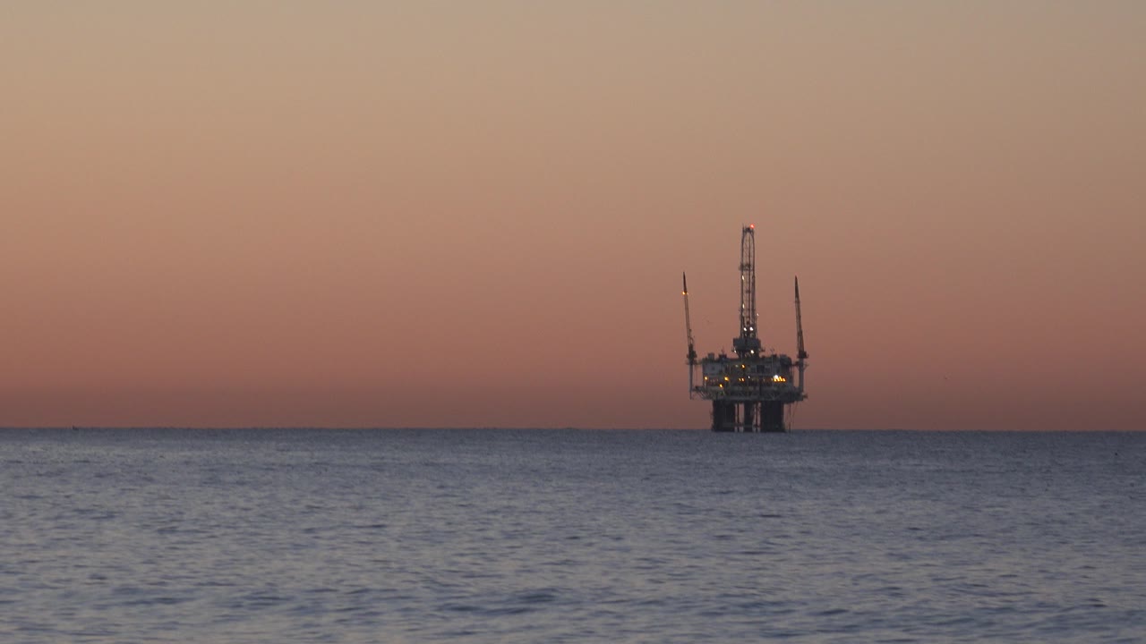 Distant offshore oil platform on the Huntington Beach ocean horizon under a pink dusk sky, with rippled water in the foreground and no visible people.