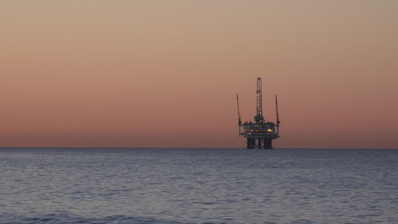 The oil rig remains centered on the distant Huntington Beach horizon, its dark structure and small lights set against pink-orange dusk with no visible people.
