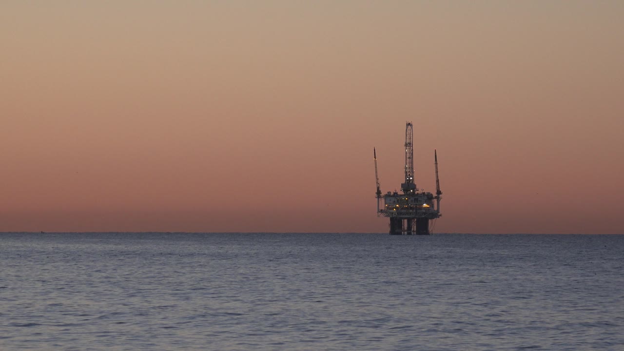 A distant offshore platform stands on the Huntington Beach ocean horizon beneath a pink-orange sunset sky, with dark water in the foreground and no visible people.