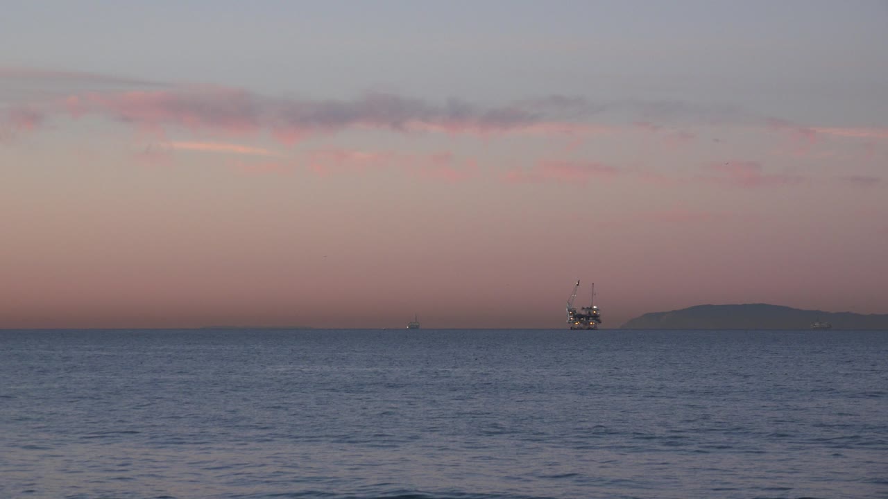 Huntington Beach ocean horizon at sundown with a small distant oil rig right of center, a dark landform behind it, soft pink sky, and no visible people.