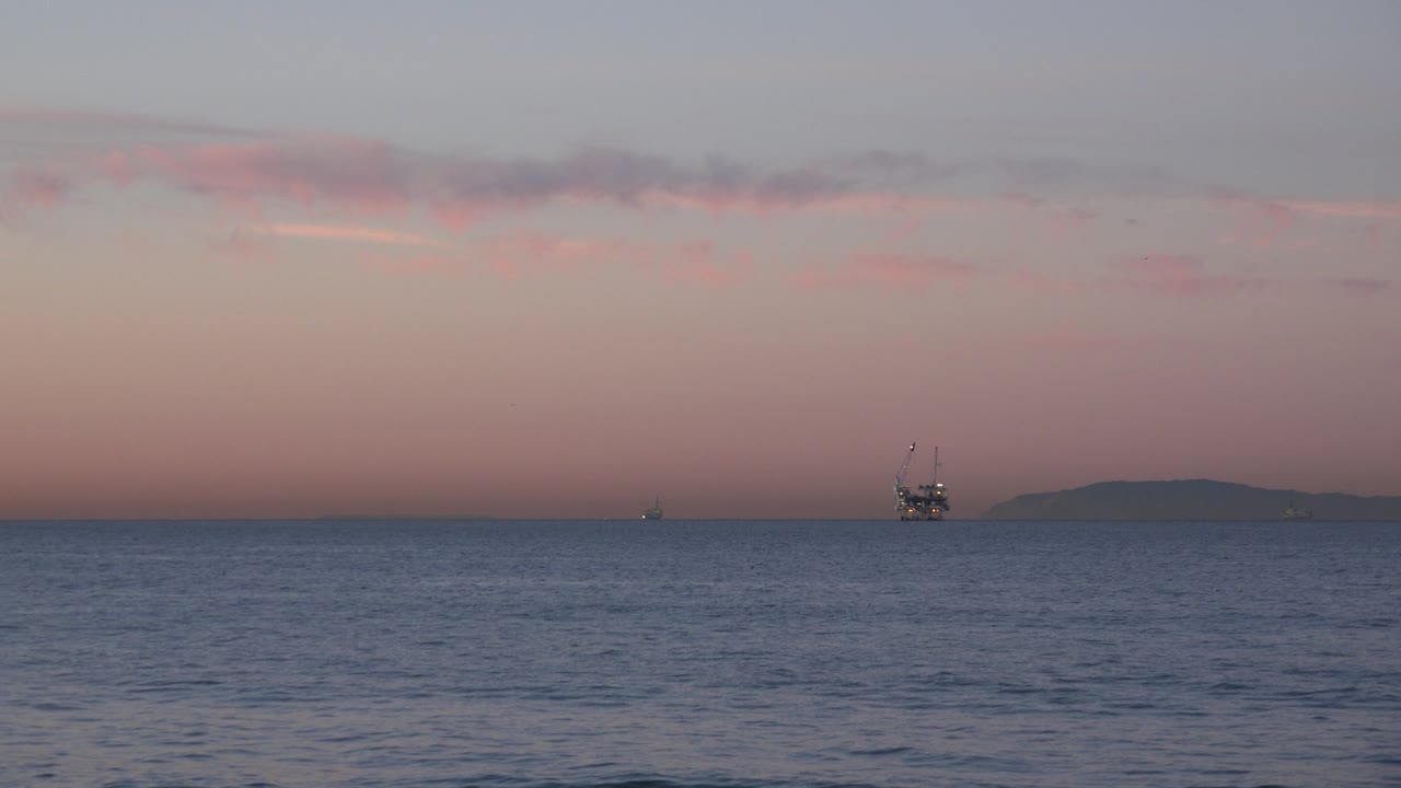 Huntington Beach ocean view with calm water in the foreground, a distant oil platform near the horizon, muted pink evening light, and no visible people.