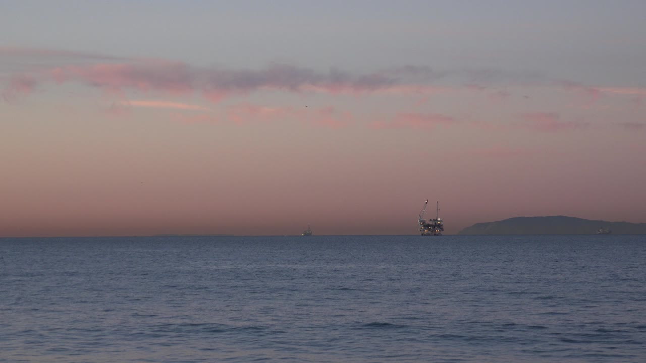 Wide Huntington Beach ocean view at dusk with a pink-orange sky, low clouds, an offshore platform near the right side, a distant landform, and no visible people.
