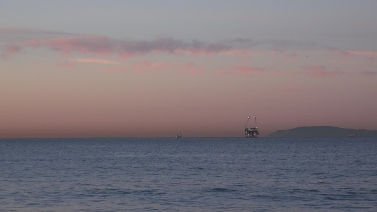 Huntington Beach water fills the foreground while a small offshore platform sits right of center beneath pink cloud bands, with no visible people.