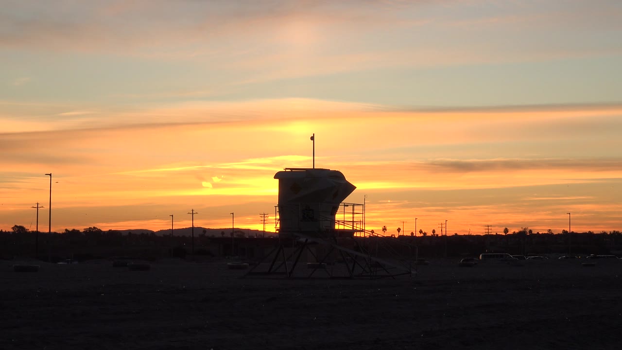 At 0:00, a silhouetted elevated structure, utility poles, and electric lines sit below an orange and pink Huntington Beach sunset sky with no visible people.