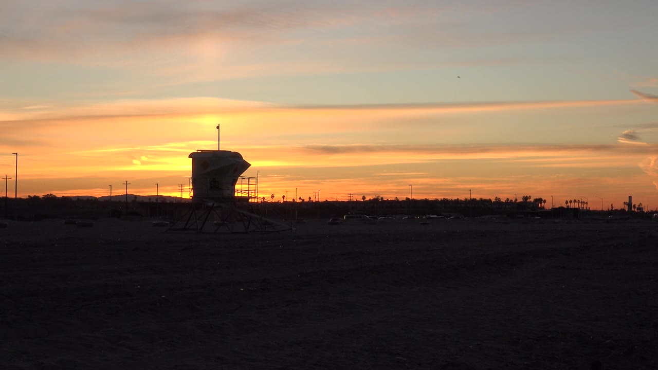 Opening frame of Huntington Beach at sunset, showing a dark roadside foreground, orange sky, silhouetted watchtower near center, distant poles, and no visible people.