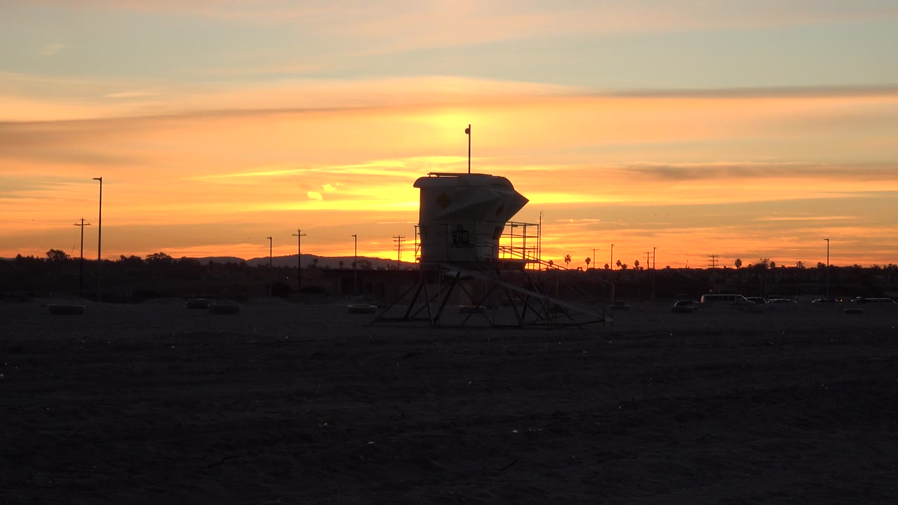 Opening frame in Huntington Beach, California, with a dark beach foreground, a silhouetted lifeguard tower near center, orange sunset bands, distant poles, and no visible people.