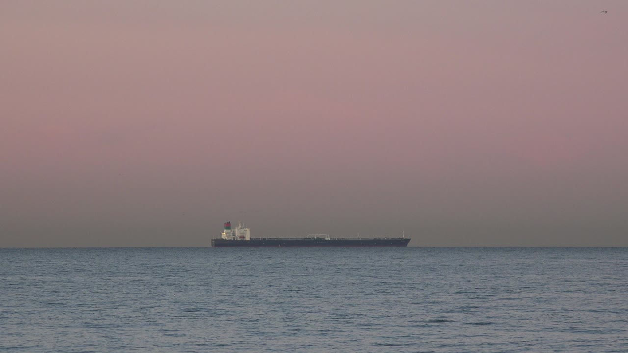 A distant ship sits left of center on the Huntington Beach ocean horizon, with low blue waves in front, pink gray sky above, and no visible people.