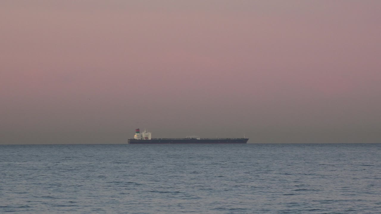 A far offshore ship sits on the right side of the Huntington Beach horizon, with muted sunset color in the sky and no visible people in frame.