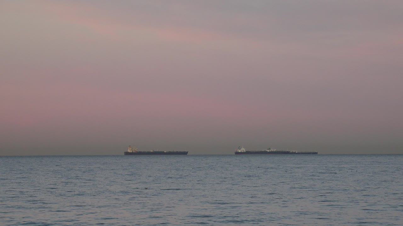 Two distant tanker ships line the horizon off Huntington Beach, California, with rippled ocean water in the foreground and no visible people.