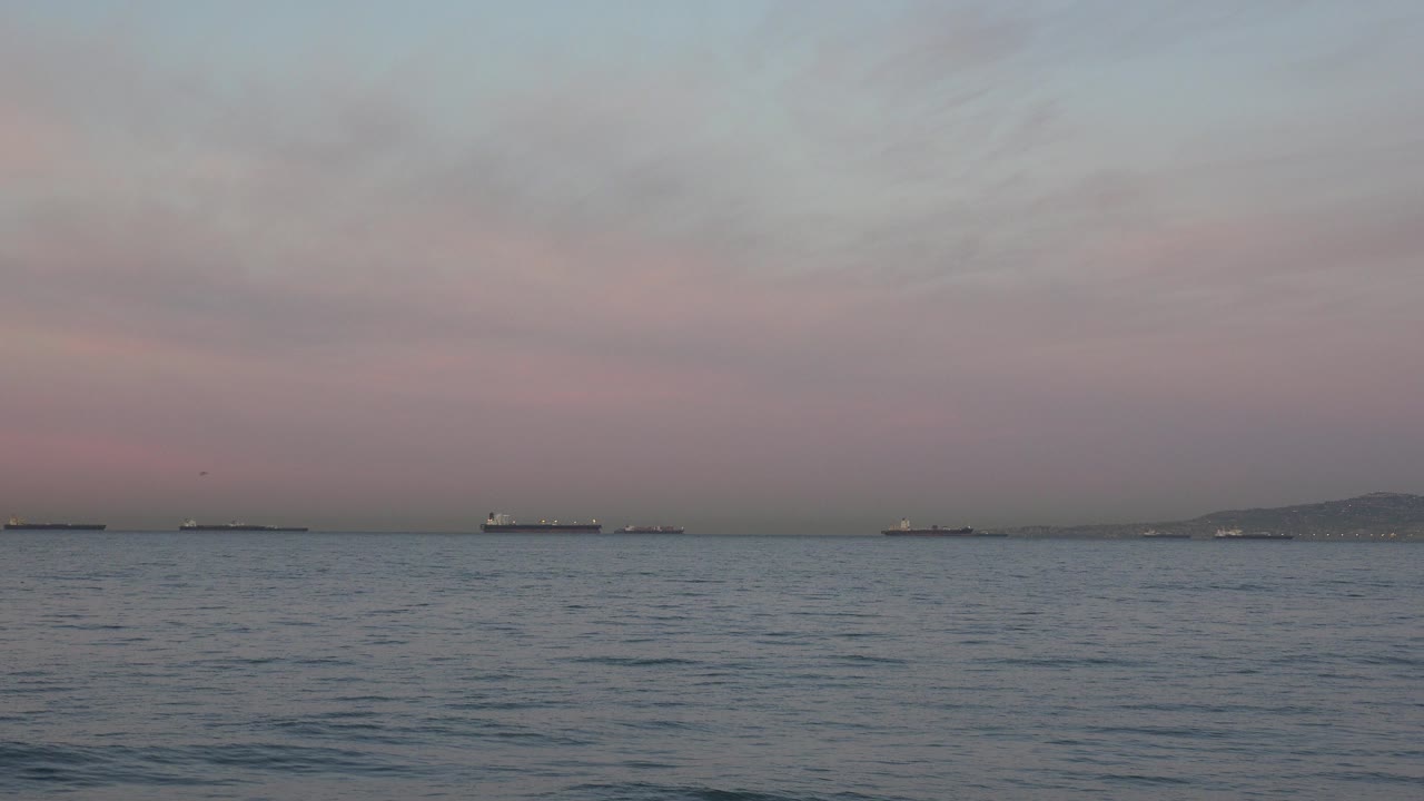At Huntington Beach, California, distant oil tankers line the horizon beyond rippled ocean water under a pale pink and gray sky, with no visible people.