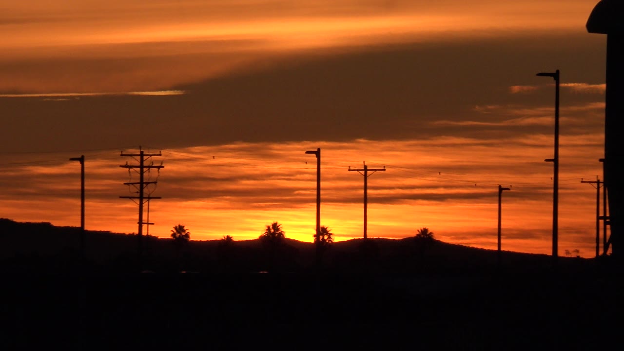 Huntington Beach sunset frame with orange layered clouds, a dark horizon, utility pole silhouettes, streetlights, sparse trees, and no visible people.