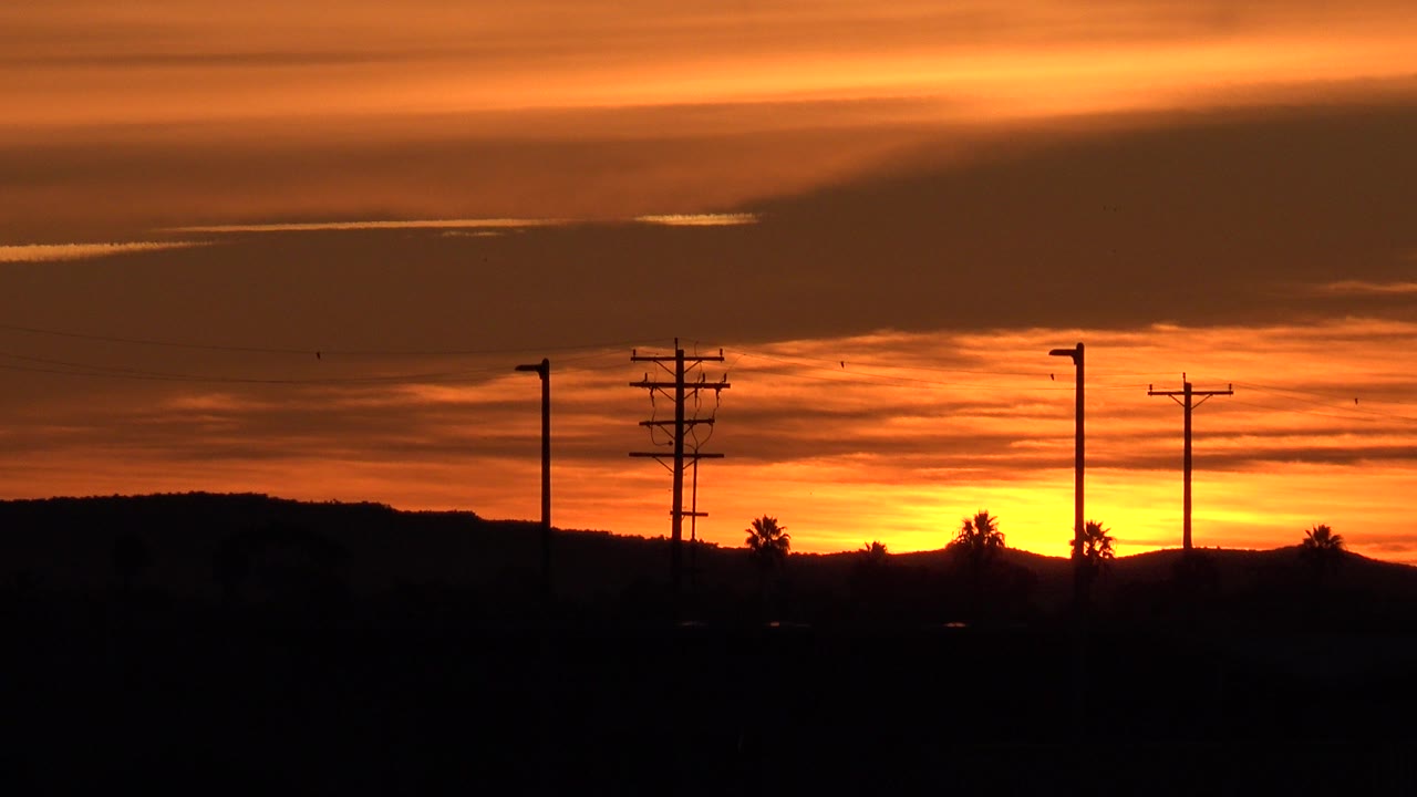 Huntington Beach sunset sky glows orange behind silhouetted utility poles, streetlights, low trees, and a dark foreground with no visible people.