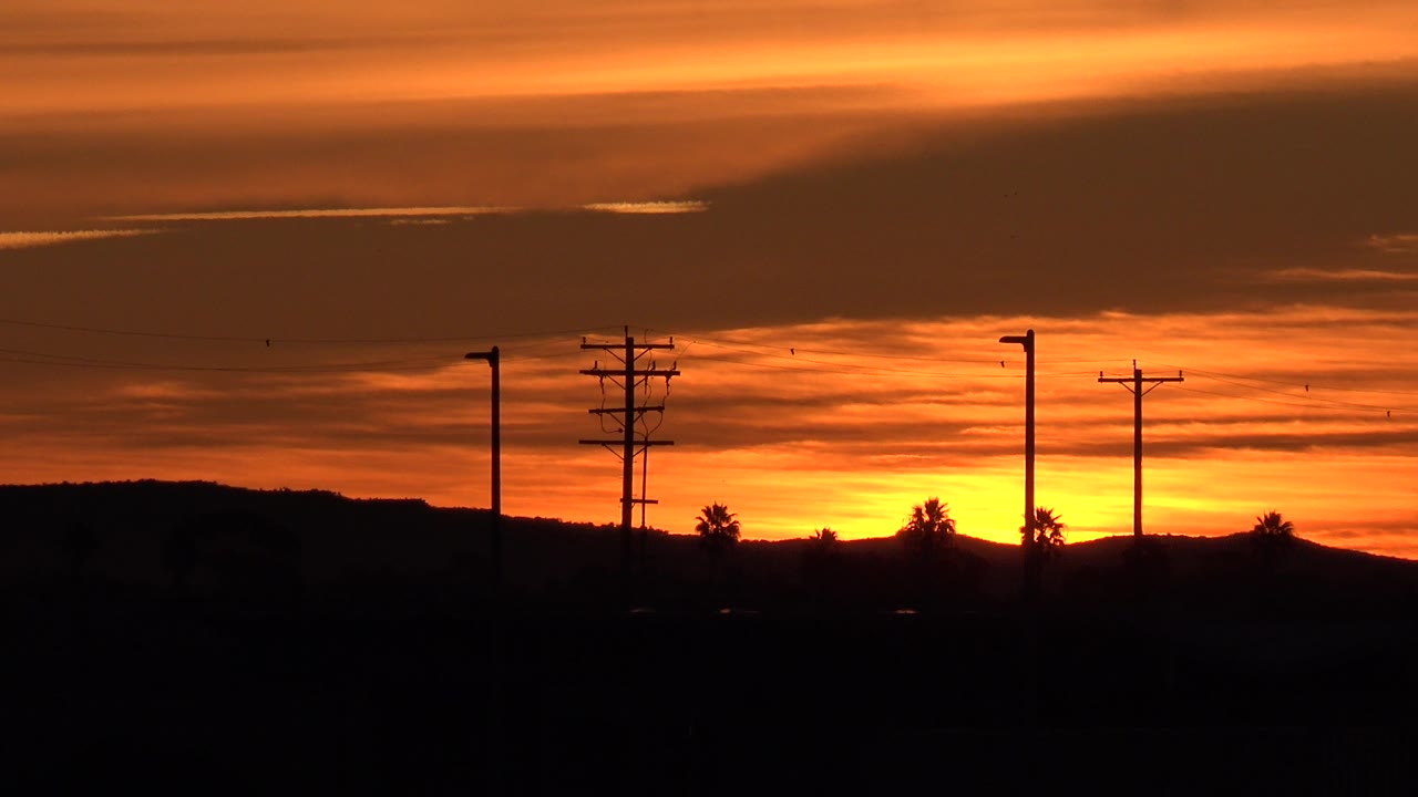 Sunset cloud layers over a dark shoreline with several pole silhouettes.