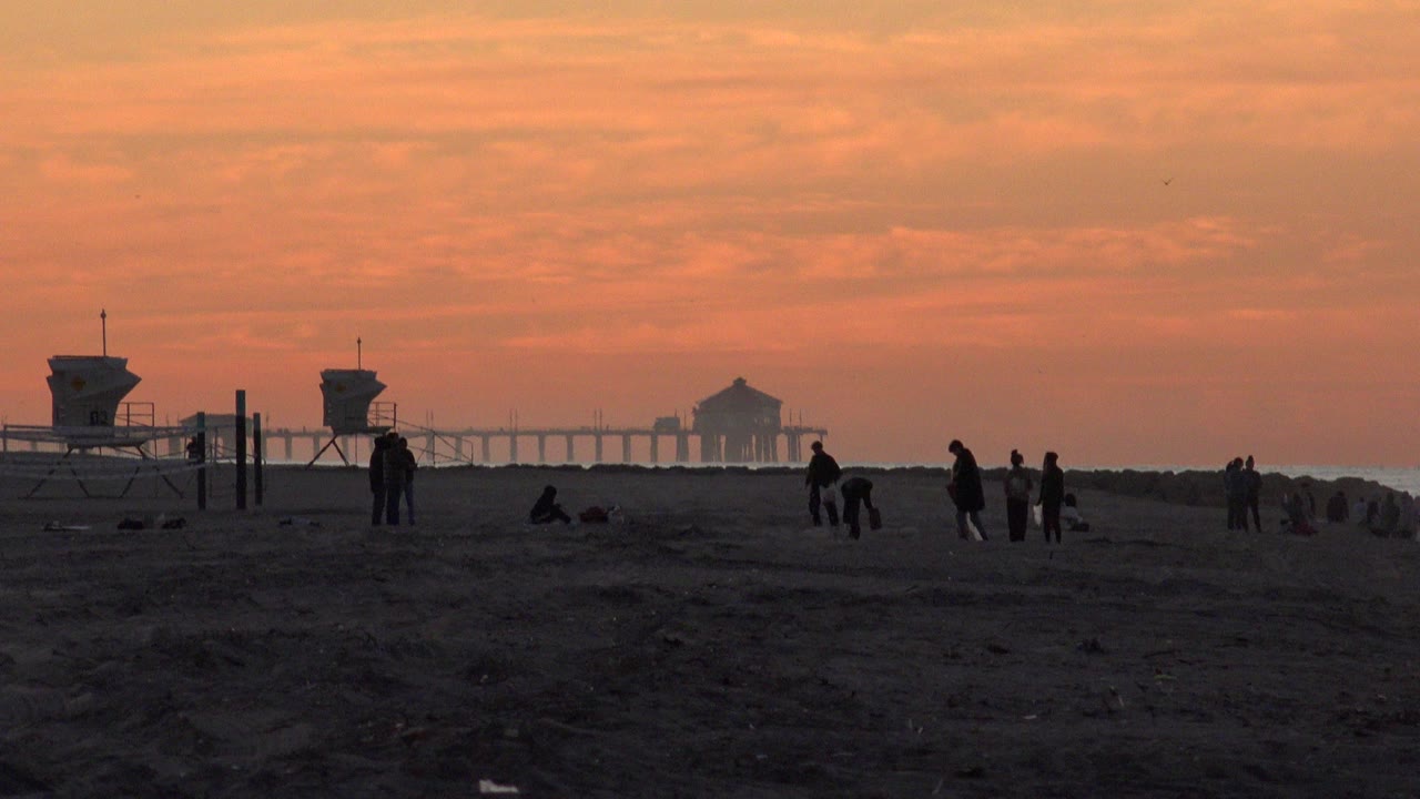 Wide Huntington Beach sunset frame with dark sand in front, scattered silhouetted beachgoers, lifeguard towers, and a pier shape on the horizon.
