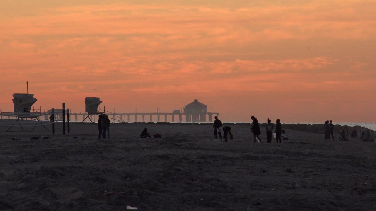 Huntington Beach view one second in, with a central lifeguard tower, people standing and sitting on the sand, and the pier faint behind the horizon haze.