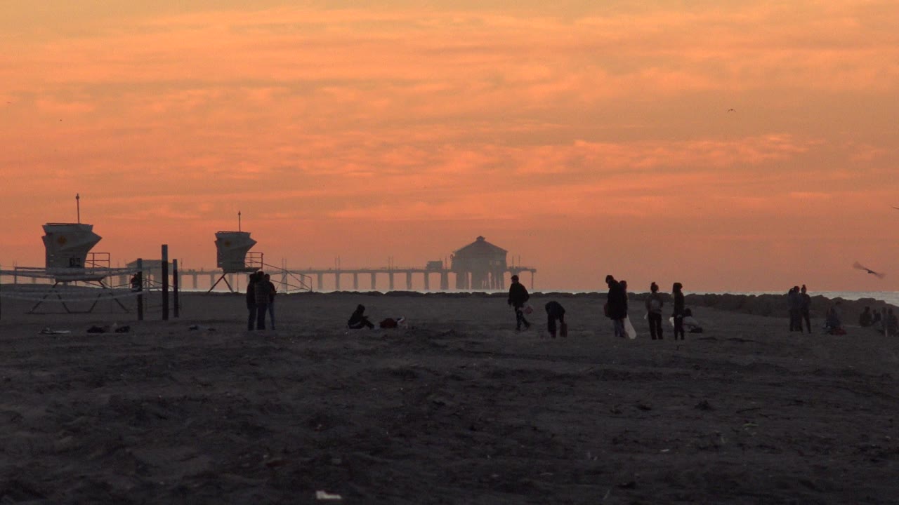 Silhouetted groups on a beach with lifeguard towers and a pier at sunset.