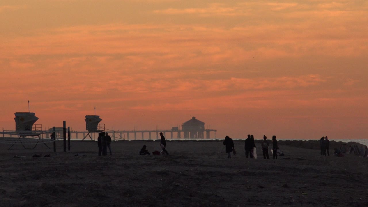 Silhouetted people on a beach at orange sunset with lifeguard towers and a distant pier-like structure.