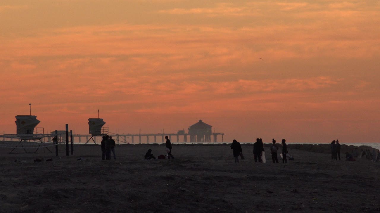 Huntington Beach, California sunset scene with a person walking near center, seated figures on the sand, lifeguard towers to the left, and the distant pier-like structure beyond the surf line.