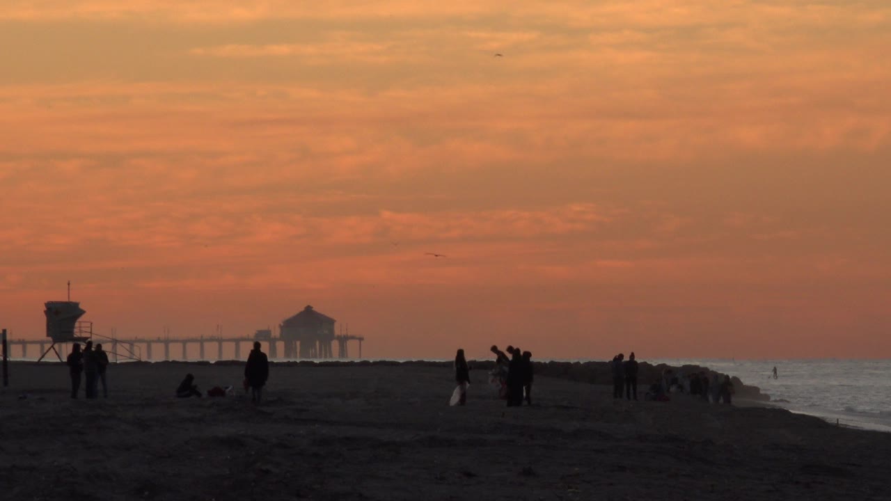 Beachgoers silhouetted on sand at sunset near a lifeguard tower and distant pier.