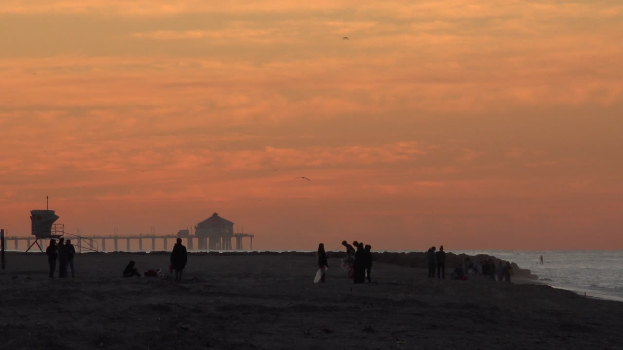 Huntington Beach frame with several silhouetted beachgoers standing and walking on the sand, the lifeguard tower left of center, a hazy pier in the distance, and water reflecting sunset light.